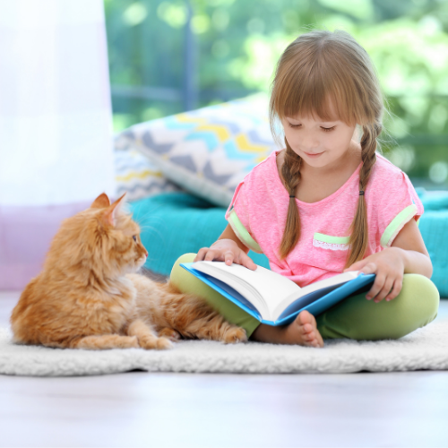 a young girl with braids wearing a pink shirt is reading to a yellow longhair cat