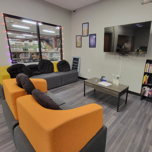 photo of cover of the teen lounge with lights in the window, gray and oranges couches facing a TV with coffee table setting